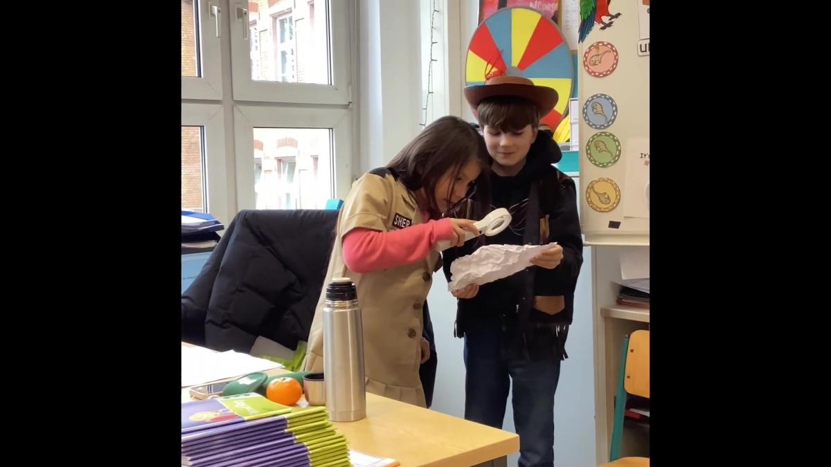   Zwei Kinder in Kostümen stehen in einem Klassenzimmer, eines trägt einen Cowboyhut, das andere untersucht ein Papier mit einer Lupe. 