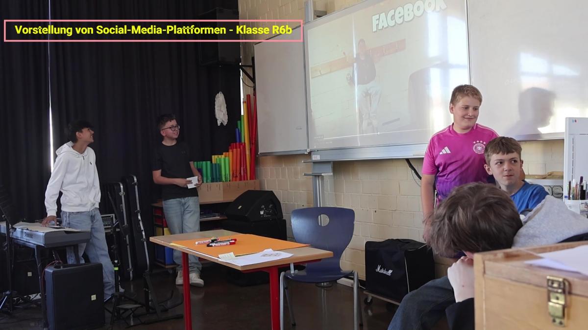 Vier Schüler präsentieren in einem Klassenzimmer. Einer steht neben einer Tafel, auf die eine Facebook-Folie projiziert wird. Der Text darüber lautet: Vorstellung von Social-Media-Plattformen - Klasse R6b